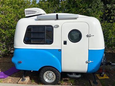 A small happier camper fiberglass trailer that is white on the top half and bright blue on the bottom half with some green bushes in the back