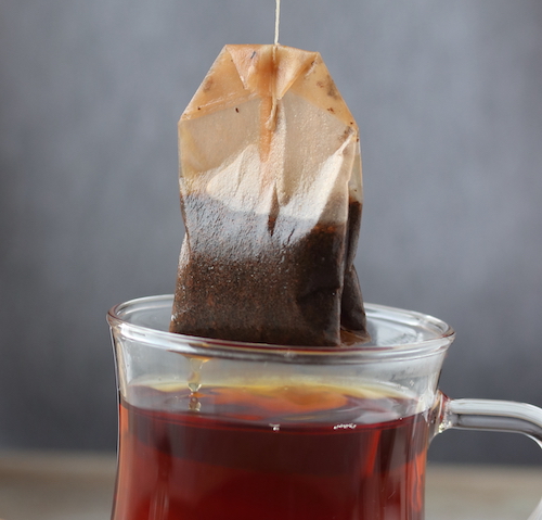 a tea bag being pulled out of a transparent mug of black tea