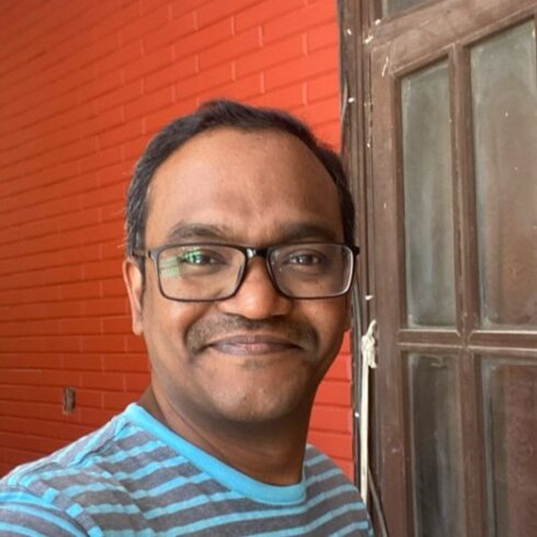 a headshot of Dr Manjunath of him smiling on a background with bright red walls and a door